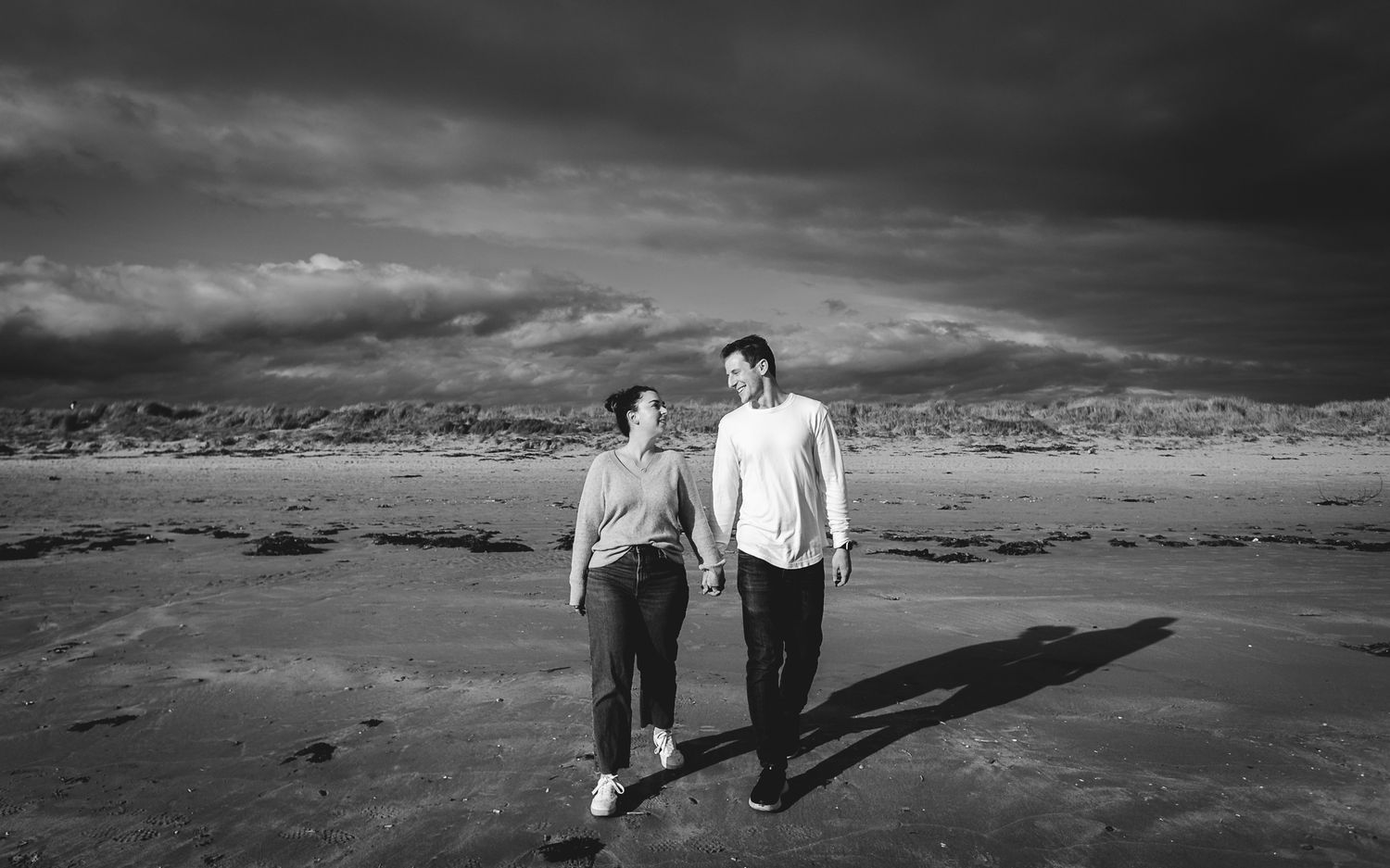 A couple walks along a dramatic black and white beach landscape with mountains and stormy clouds overhead.