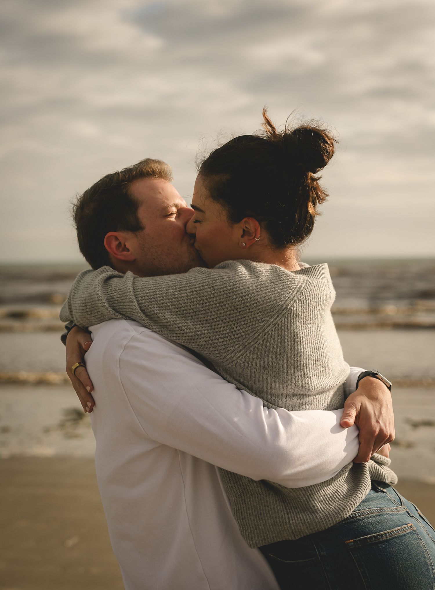 A romantic couple sharing an intimate kiss on a beach at sunset while embracing in casual sweater and jeans.