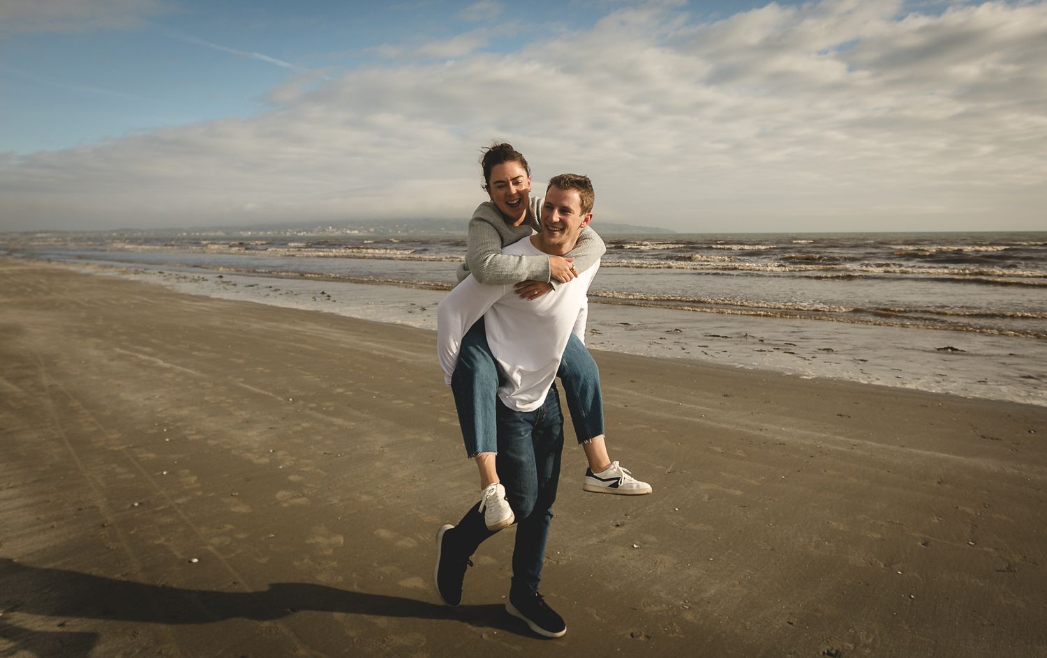 A couple shares joyful moments on a serene beach with dramatic cloudy skies during golden hour.