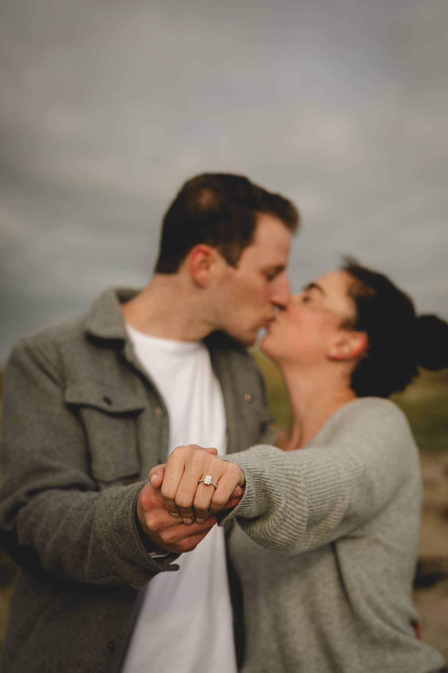 A couple shares a romantic moment outdoors while clasping hands, with one wearing grey and the other in white.