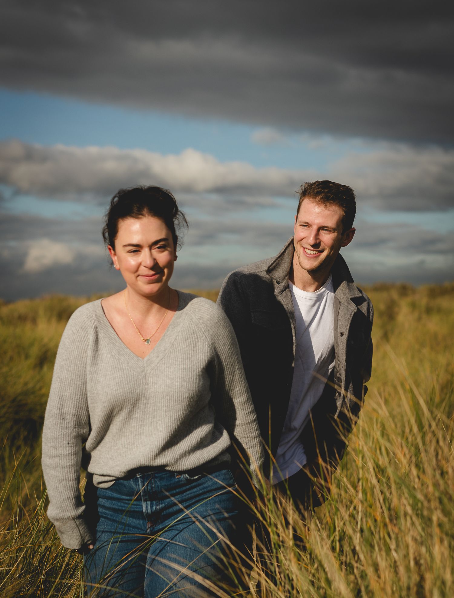 Couple in casual attire sitting together in tall golden grass during sunset with dramatic cloudy sky.