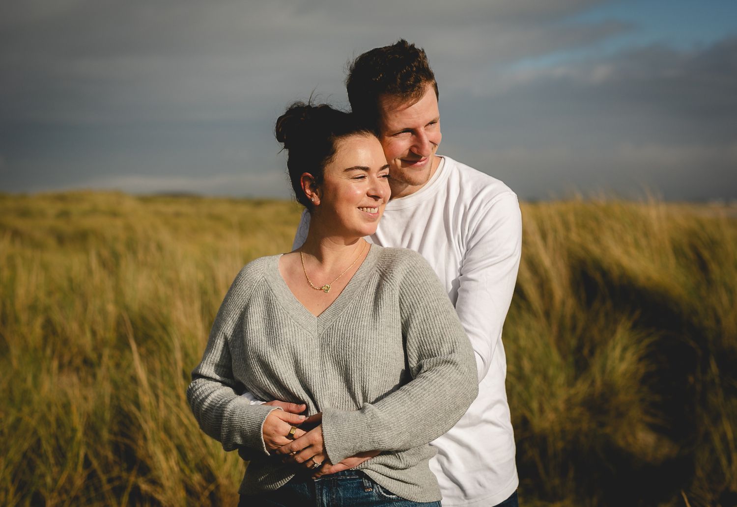 A couple embraces in a golden wheat field at sunset under dramatic cloudy skies.