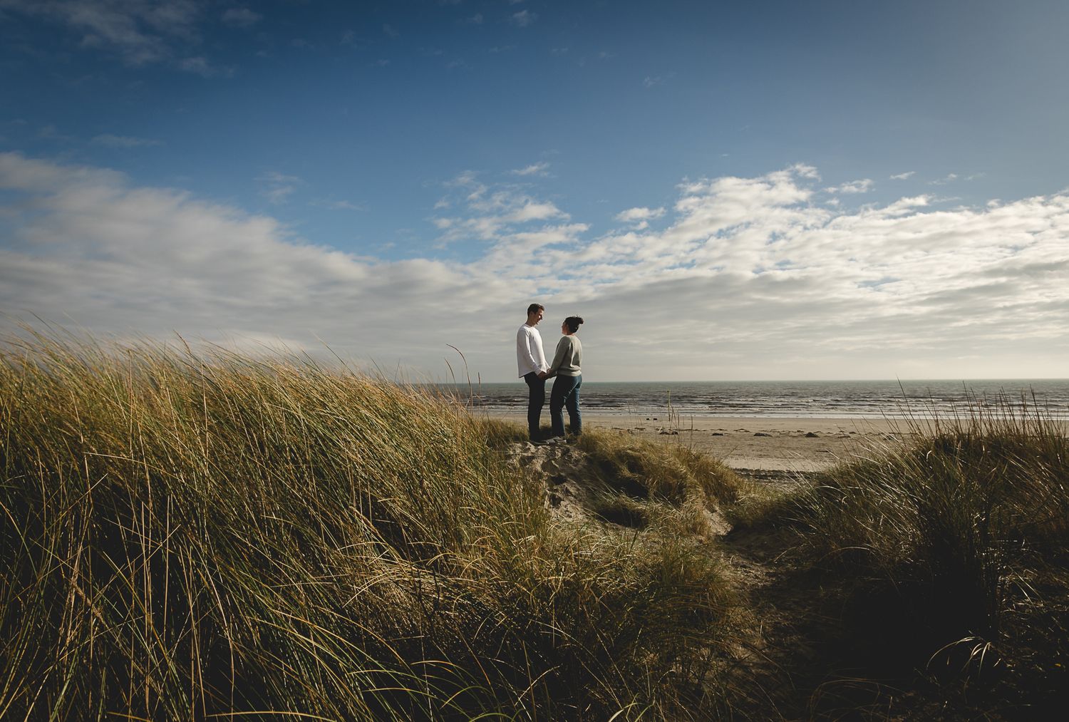 Two figures stand together on coastal dunes with tall grass and cloudy skies at sunset.