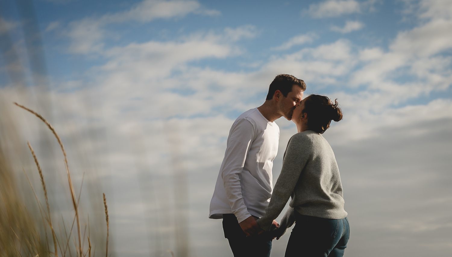 A romantic couple stands close together among beach grass at sunset with cloudy sky in the background.