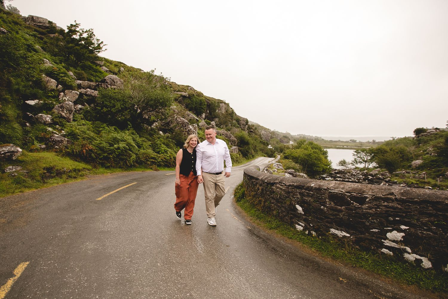 A couple walks together on a winding coastal road surrounded by green hills and rocky landscapes in Ireland.