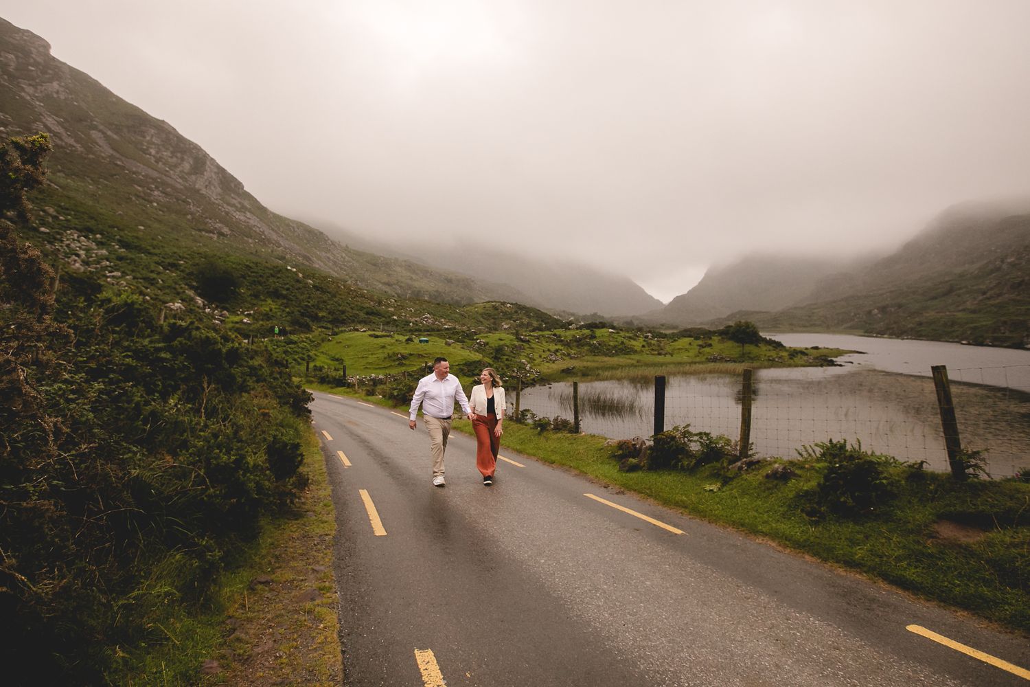 Misty mountain landscape with a winding road through green hills and a lake in the distance.