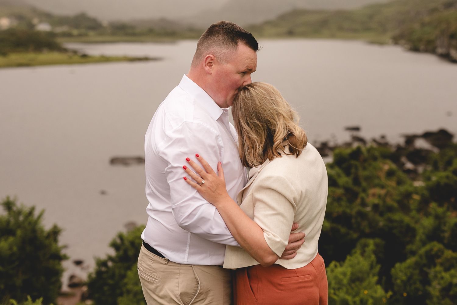 Couple embraces in a romantic moment against a misty mountainous lakeshore backdrop.