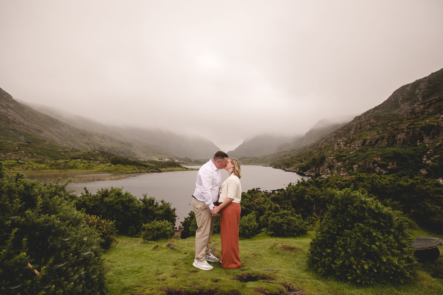 Couple shares romantic moment on grassy hilltop overlooking misty lake valley in Ireland.