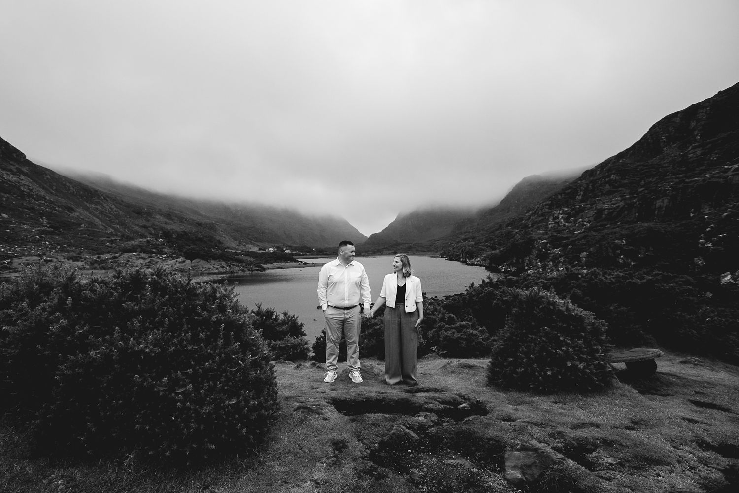 Two figures stand atop a rocky overlook with a misty lake and mountains in black and white scenery below.