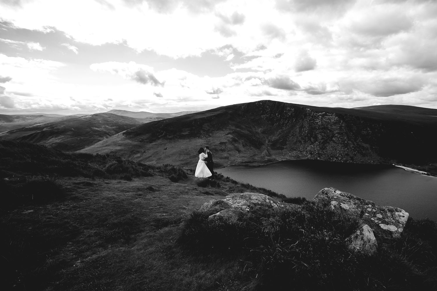 A figure sits on a rocky cliff edge overlooking a serene mountain lake under dramatic cloudy skies in black and white.