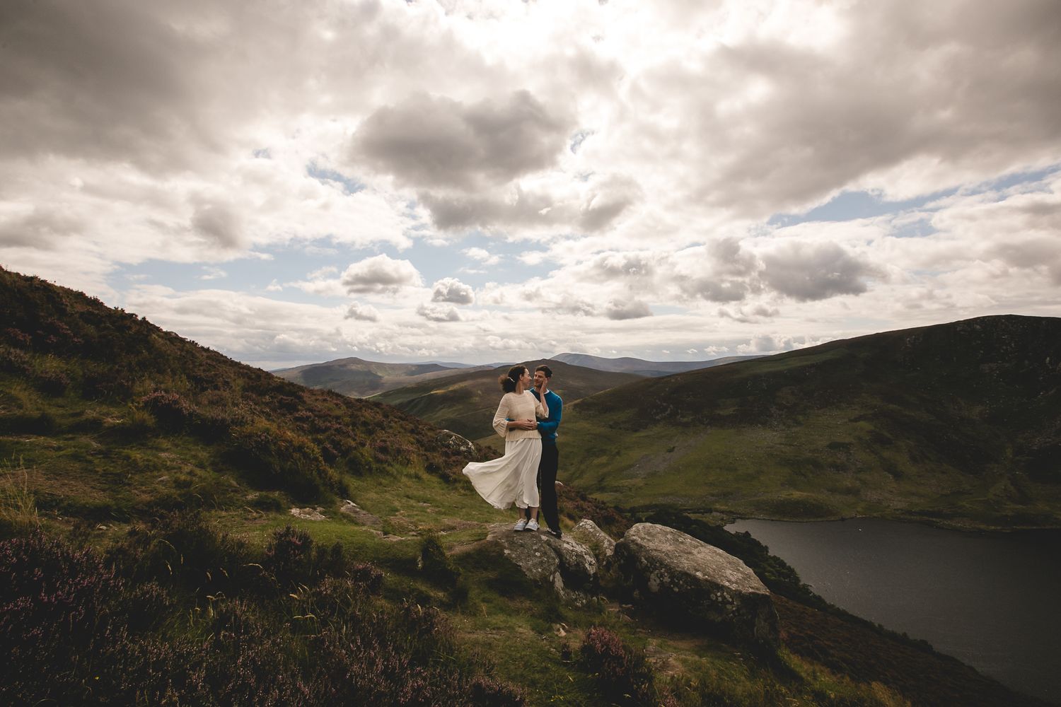 A romantic couple embraces on a dramatic hilltop with moody clouds and a serene lake valley below.