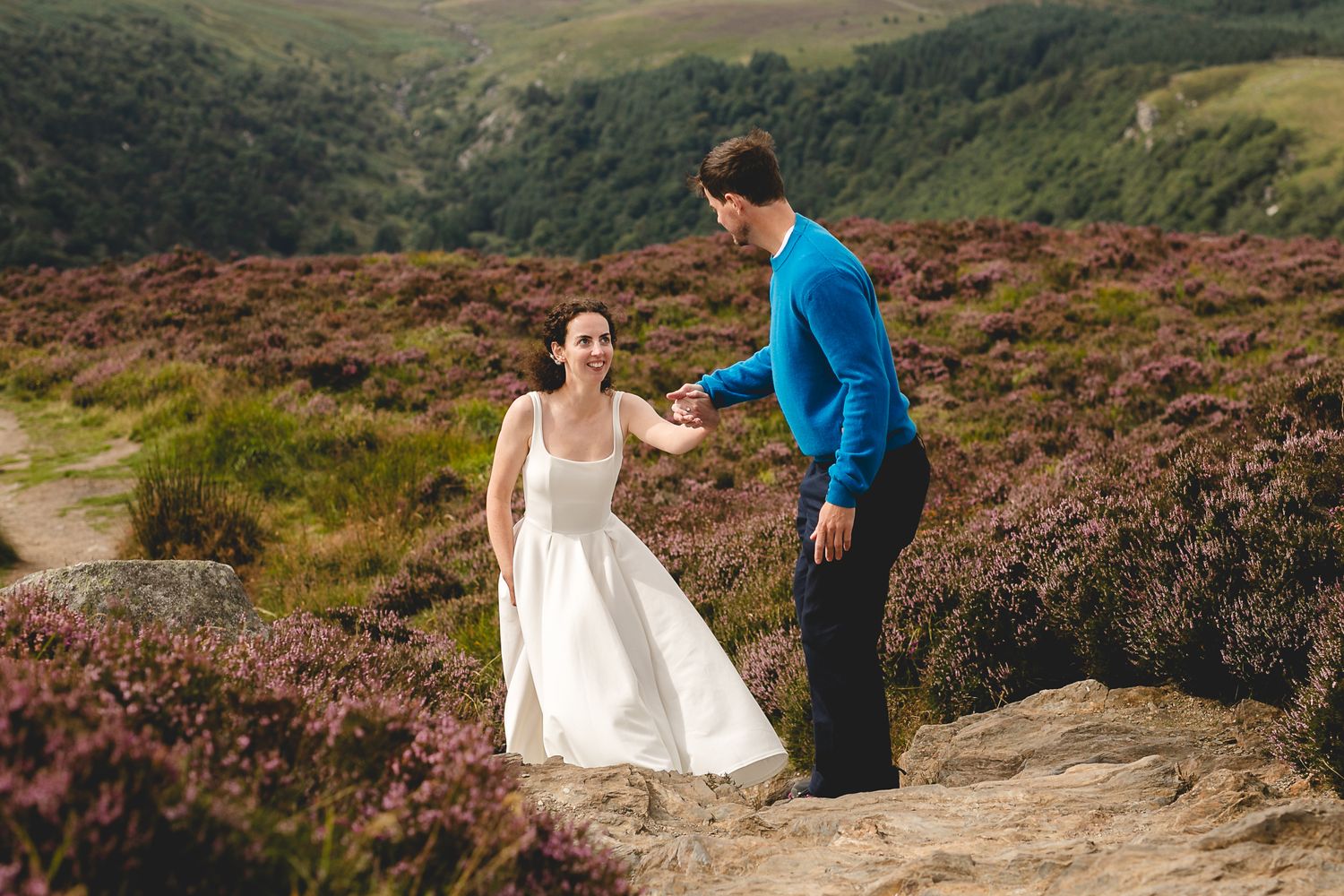 A couple in wedding attire sharing a joyful moment among purple heather on a mountainside path.