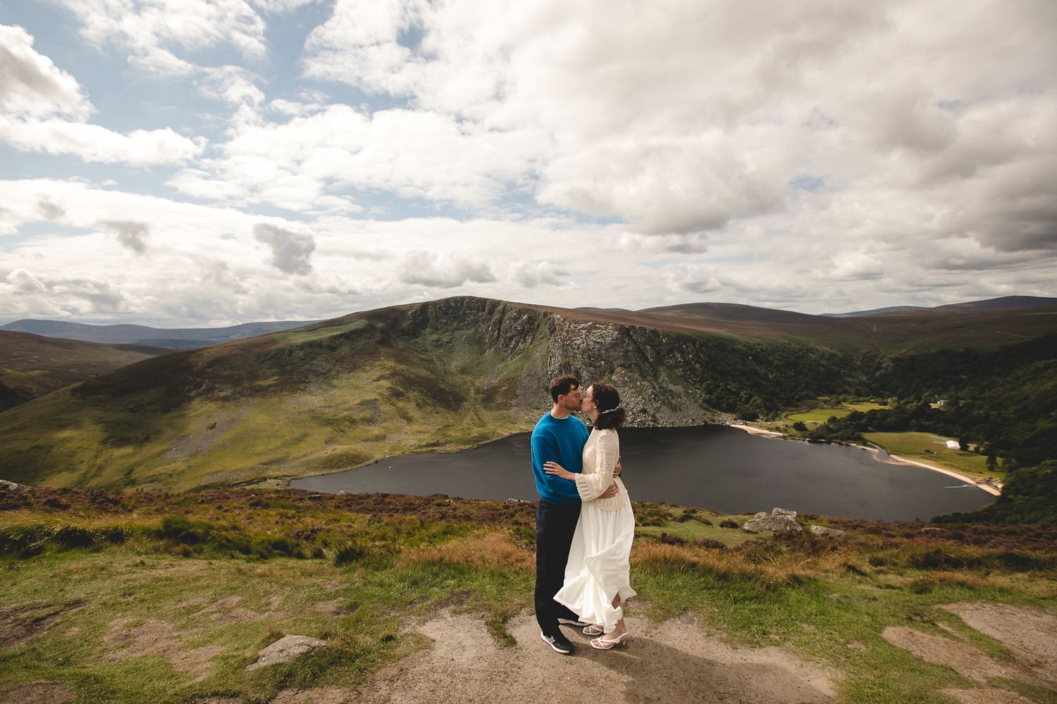 A couple embraces at a scenic mountain overlook above a lake in stunning Irish countryside under dramatic clouds.