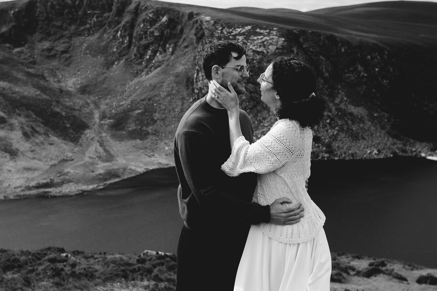 A romantic couple embraces while standing on a cliff overlooking a scenic mountain lake in black and white.