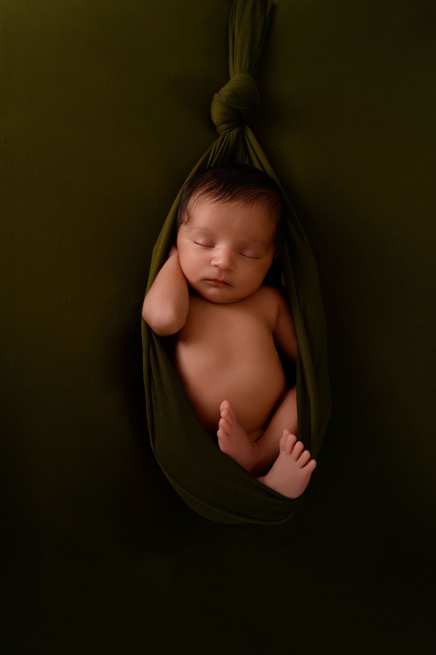 Artistic newborn portrait in dark moody lighting with baby nestled in green fabric.