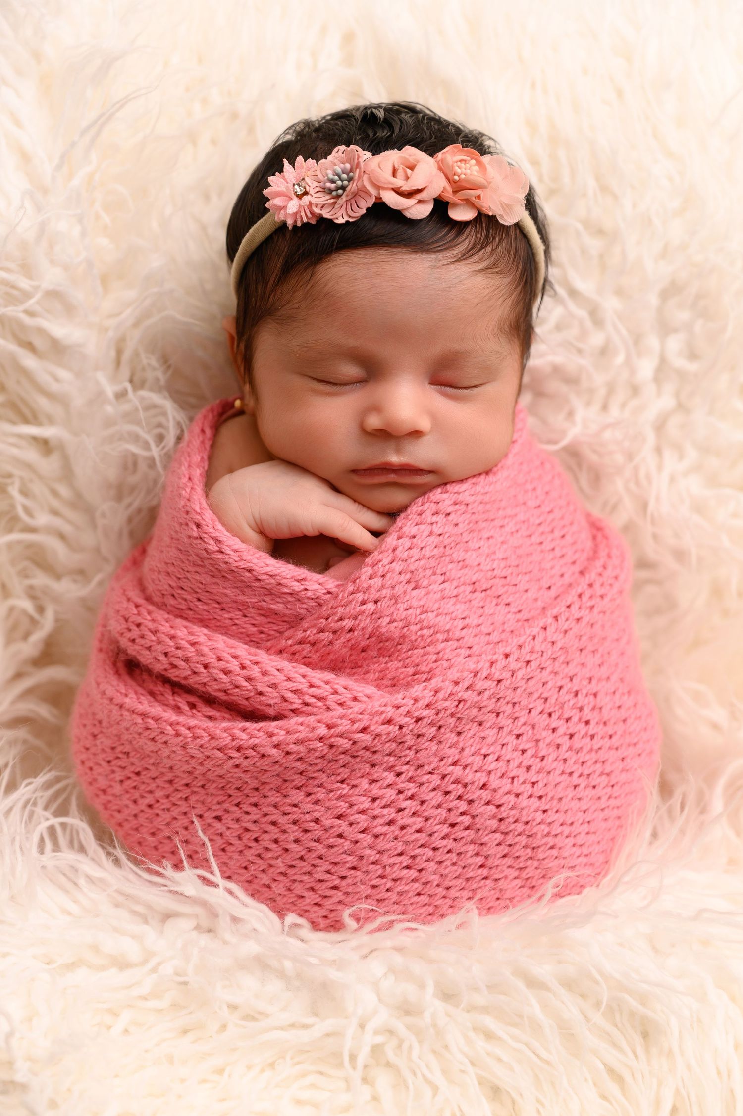 Baby sleeping peacefully in pink wrap with floral headband on soft white fur backdrop.