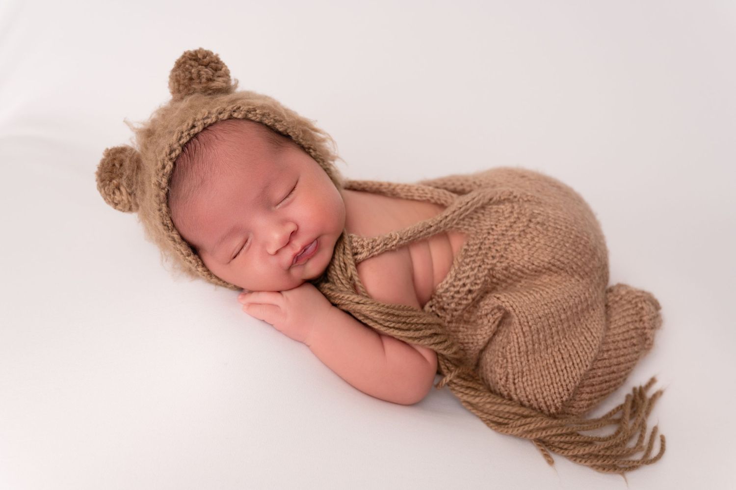 Sleeping newborn wearing knitted bear bonnet on white background in studio setting.