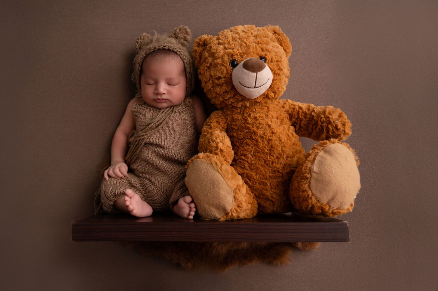 Newborn baby posed with large teddy bear on brown studio backdrop.