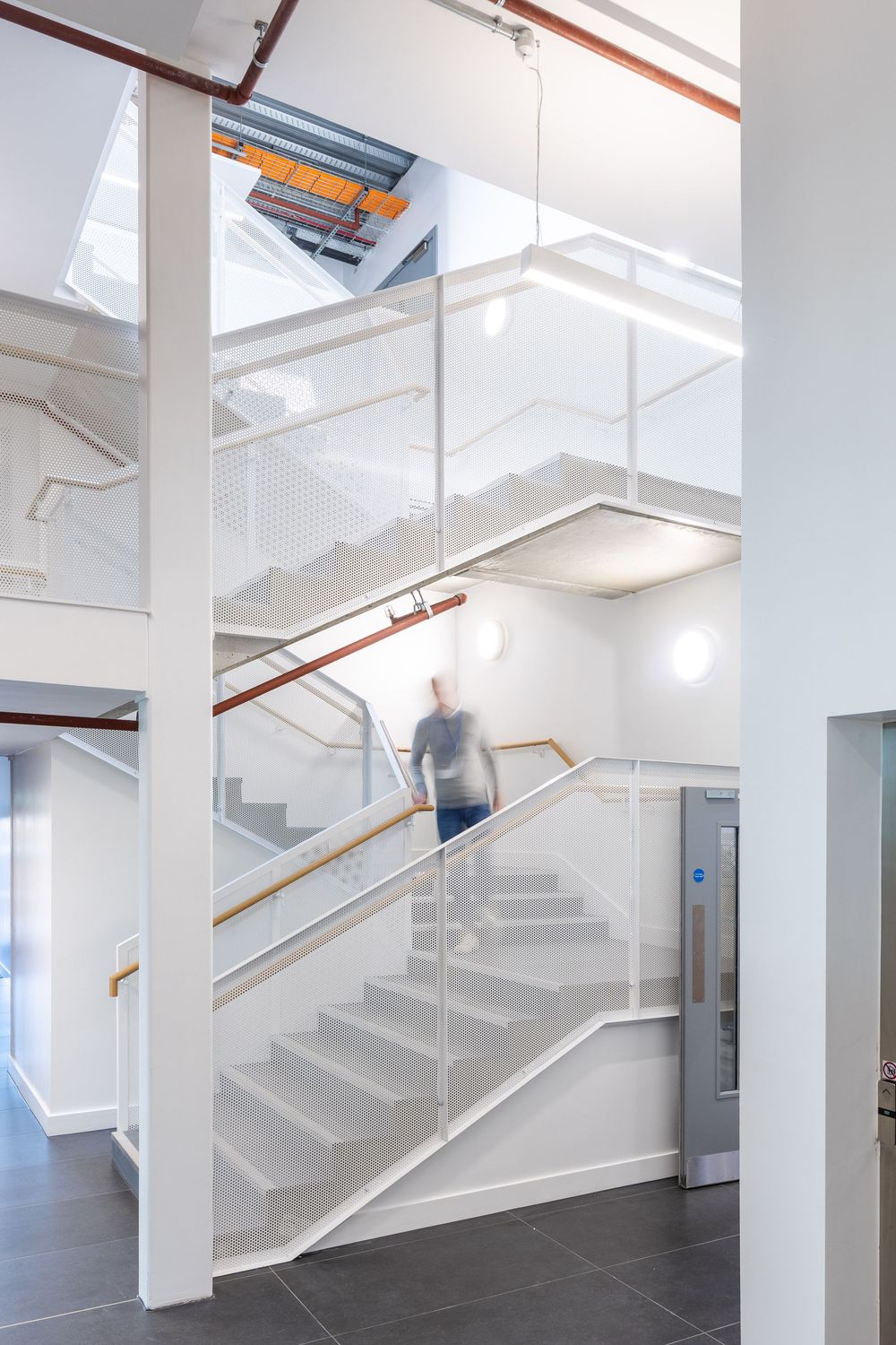 Bright modern staircase interior with perforated metal railings, clean white walls, and a blurred figure walking down the steps.