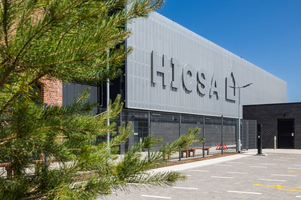 Exterior view of the HICSA building with modern metal cladding, clear signage, and greenery in the foreground under a bright blue sky.
