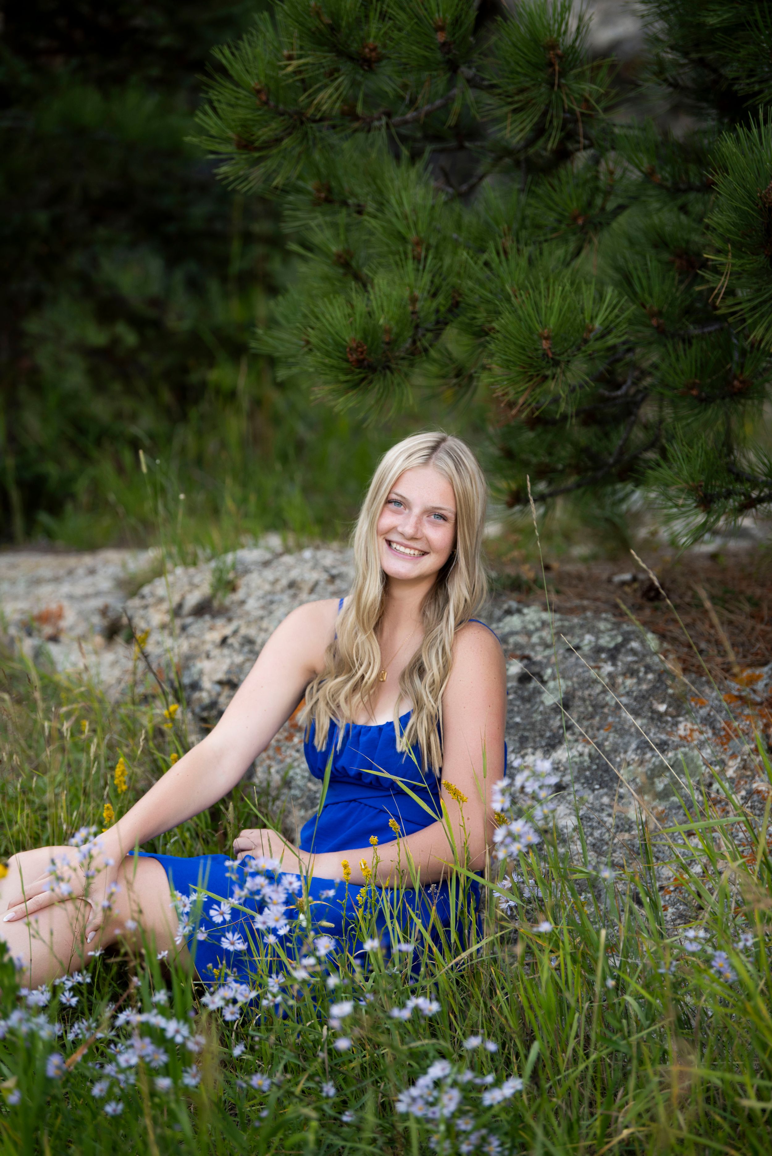Person in blue dress poses among wildflowers and pine trees in outdoor portrait session.