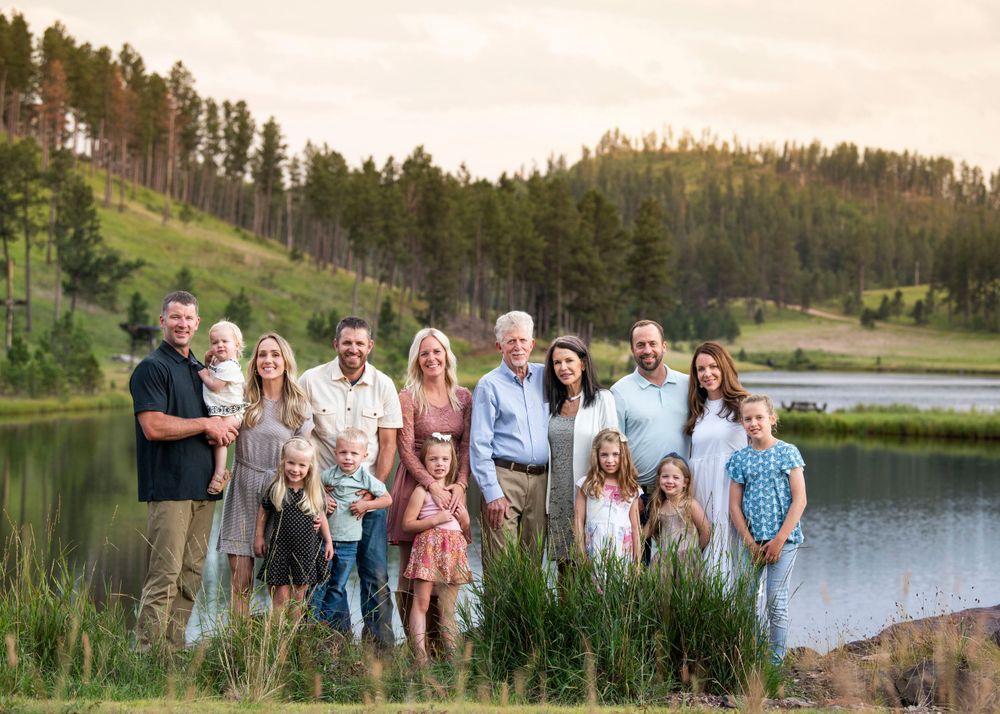 A large family group poses together by a serene lake surrounded by pine-covered hills.