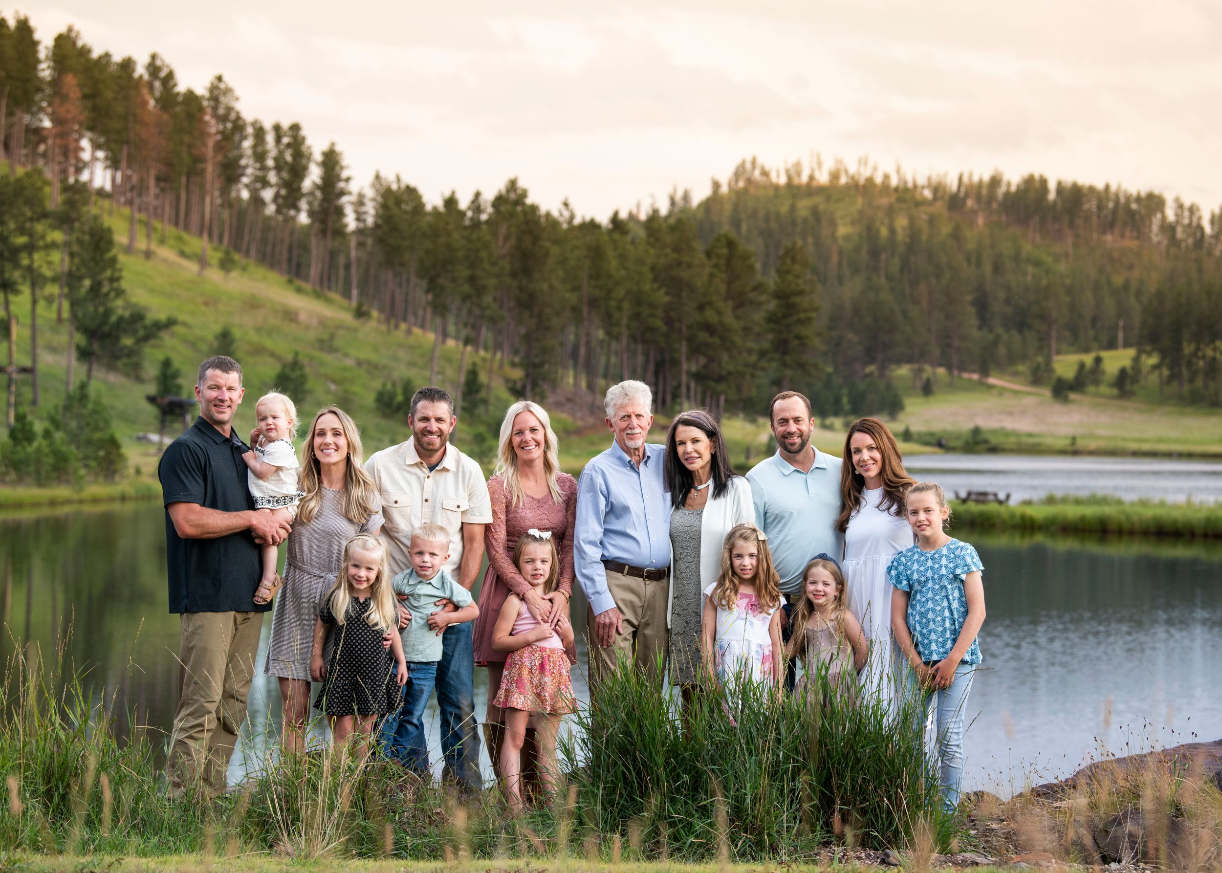 A large family group poses together by a serene lake surrounded by pine-covered hills.