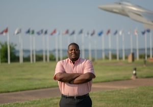 A figure stands confidently with crossed arms in front of a row of international flags and aircraft display.