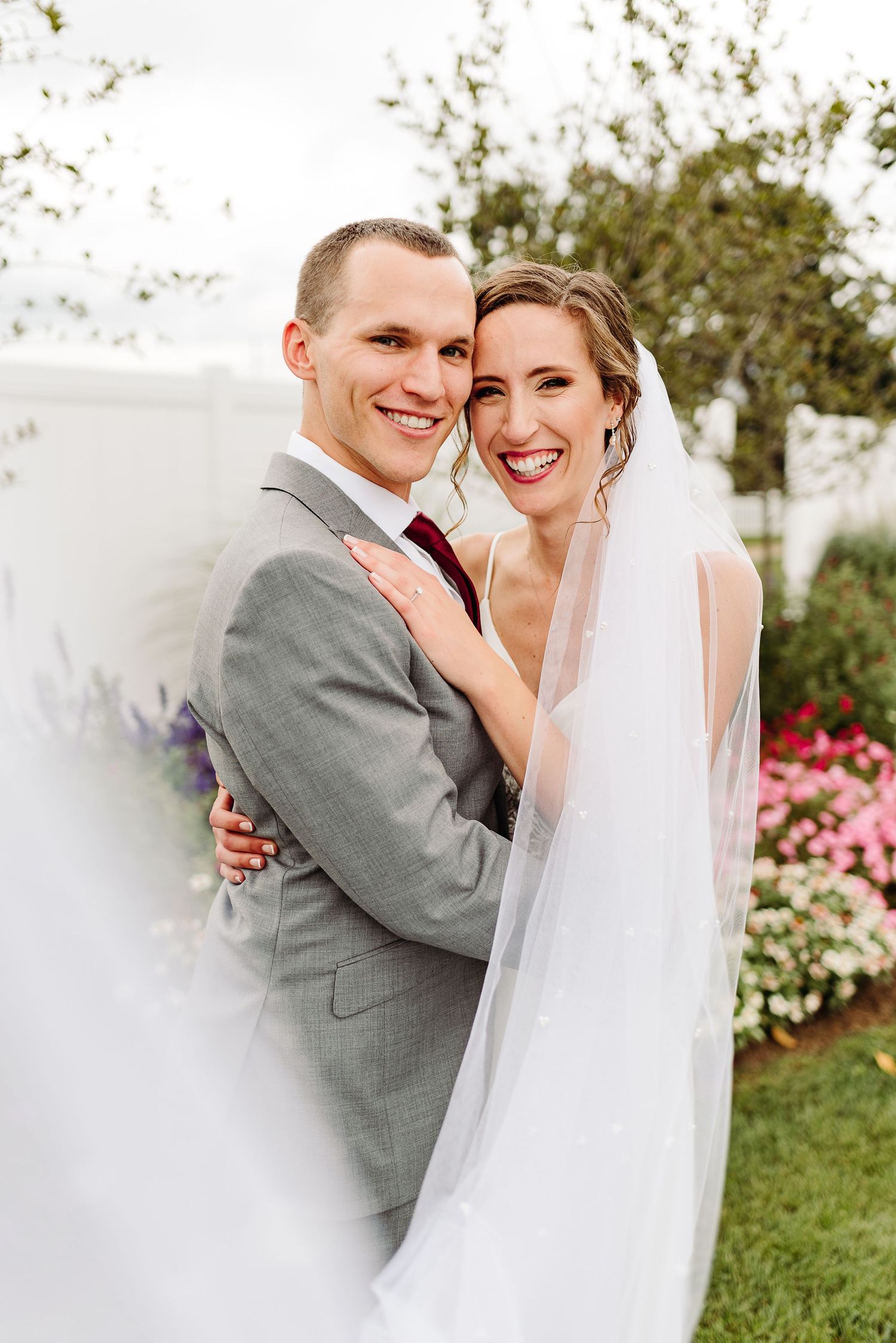 A joyful bride and groom embrace outdoors with pink flowers and greenery in the background.