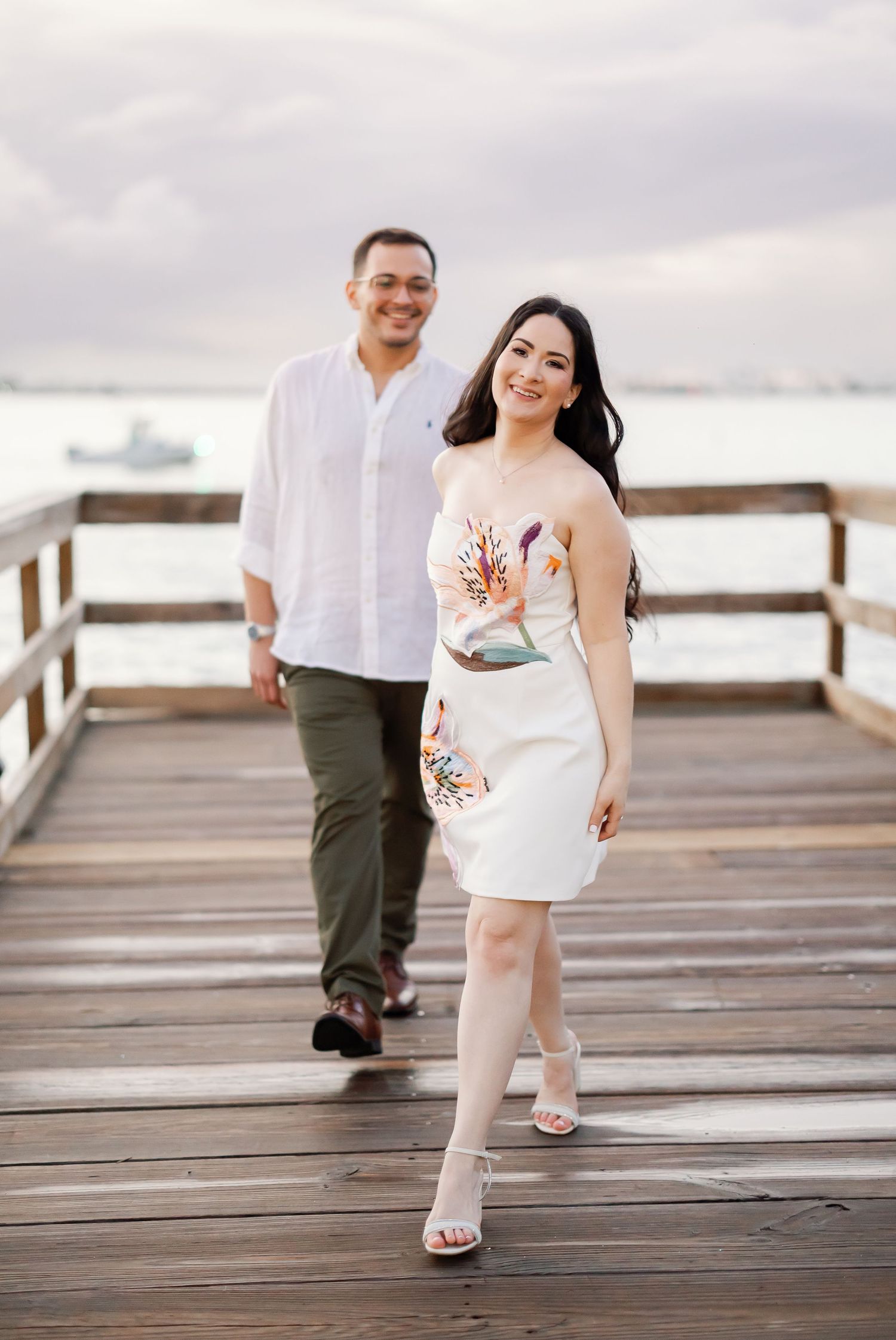 A man and a woman in casual attire stand together on a wooden pier overlooking a body of water.