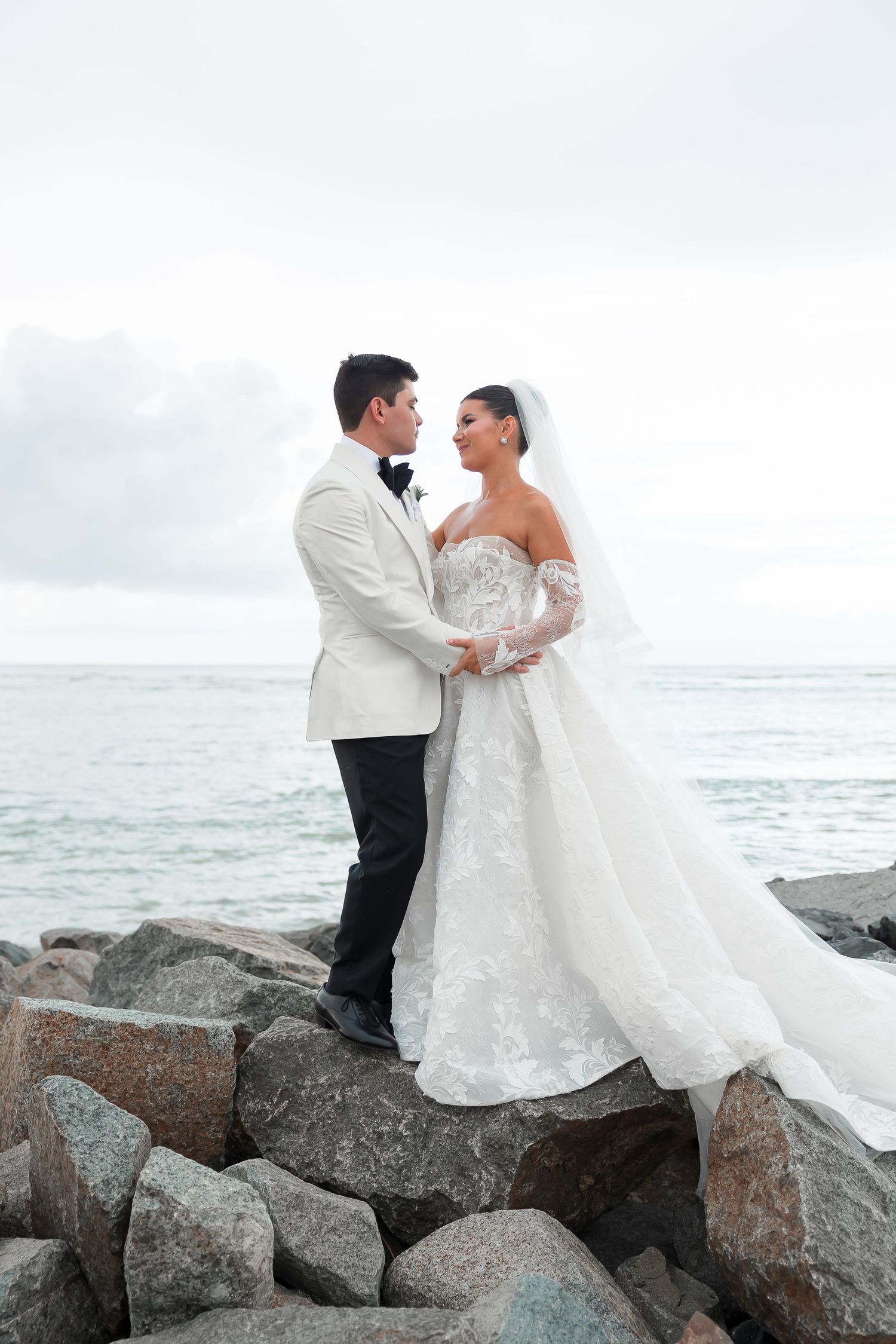 A couple hold hands on a beach, with the ocean and rocky coastline in the background.