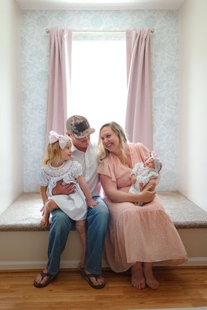 A family sits together on a window seat with pink curtains and floral wallpaper in a bright, airy room.