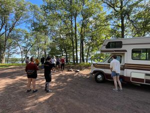 Group of people gathered around RV camper van in wooded campground area.
