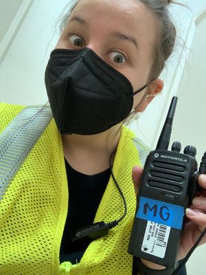 Safety worker wearing black mask and high visibility vest holds up Motorola walkie talkie radio.