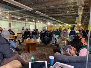 Large group meeting in industrial space with people sitting on floor around coffee table.