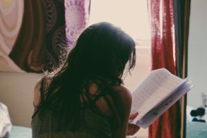 A reader silhouetted near a window with curtains studies the pages of an open book in warm lighting.