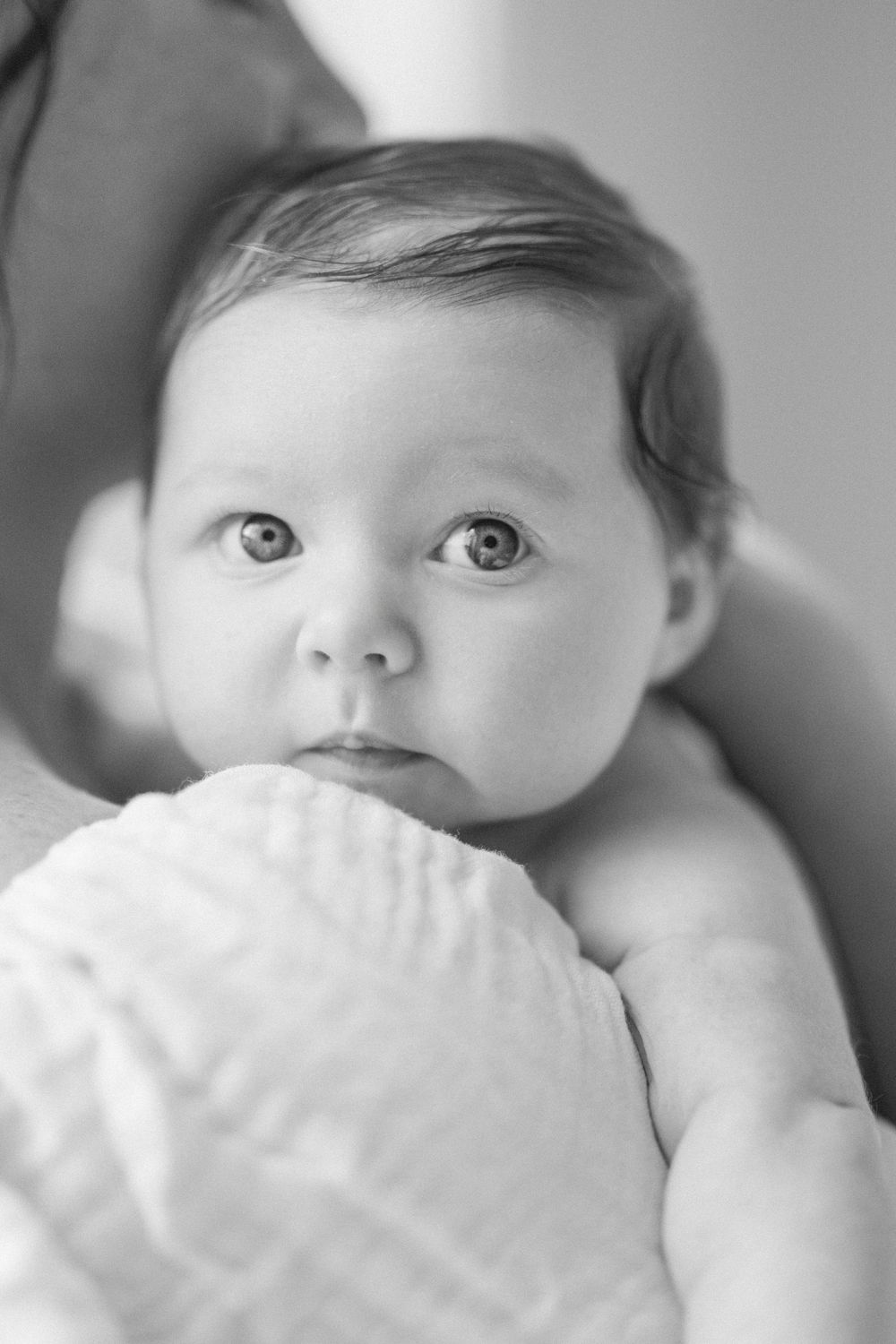 Black and white portrait of an alert newborn baby with big eyes, resting on a soft white blanket.