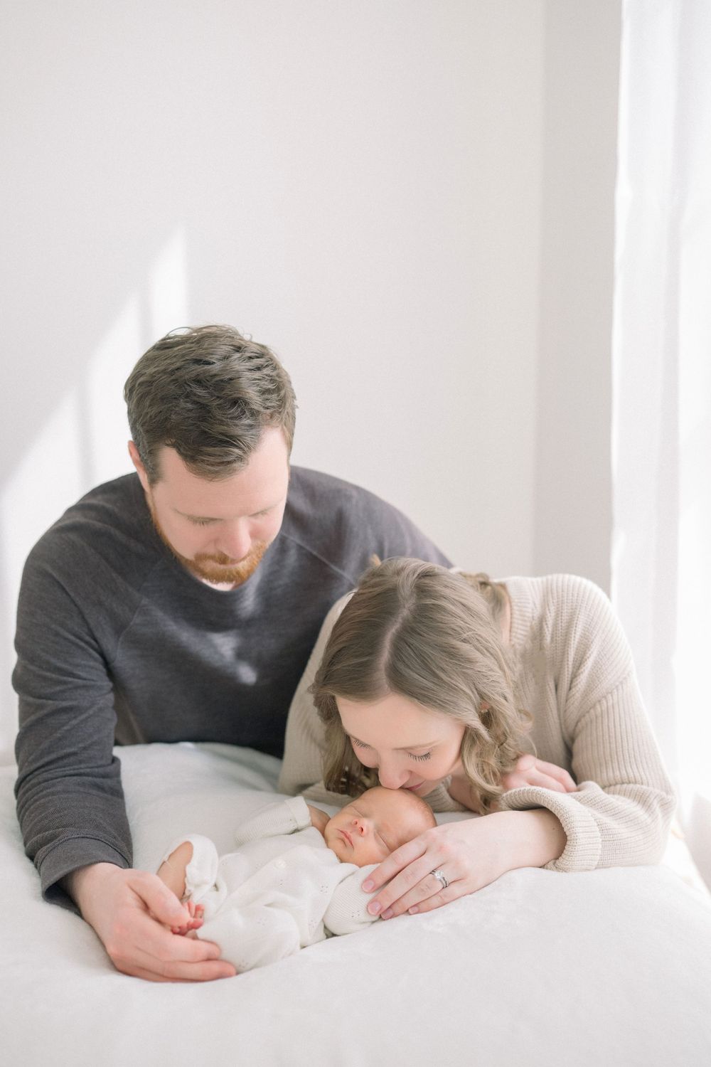 New parents lovingly gaze at their sleeping newborn baby wrapped in white during an indoor lifestyle photo session.