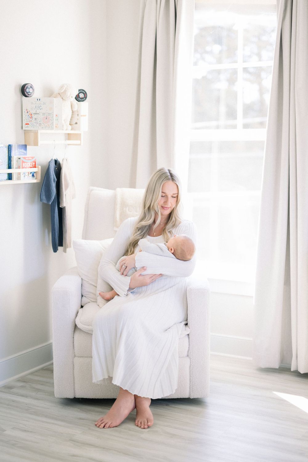 A serene mother in white dress sits in a glider chair, cradling her newborn baby in a bright, modern nursery.