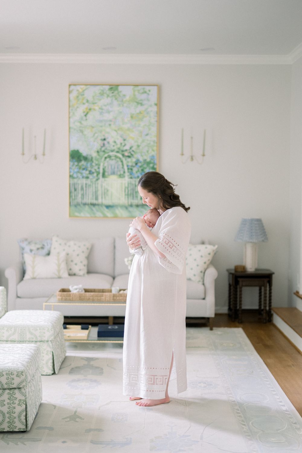 A mother in a white gown cradles her newborn in a soft, elegantly decorated living room with pastel tones.