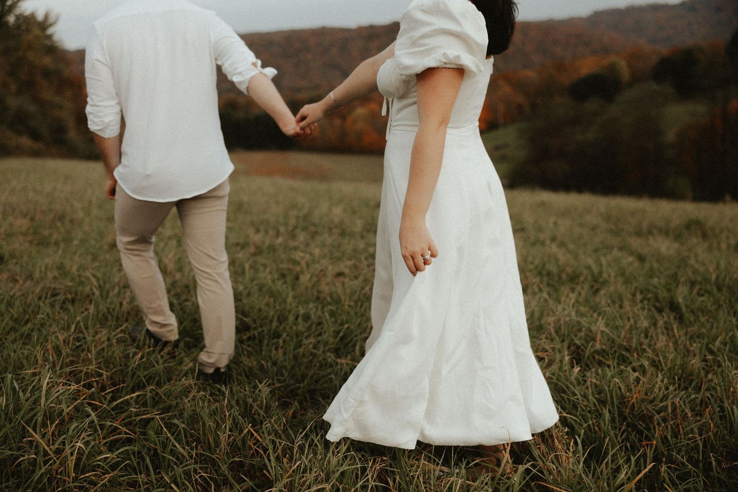 Couple holding hands while walking through grassy field at sunset.