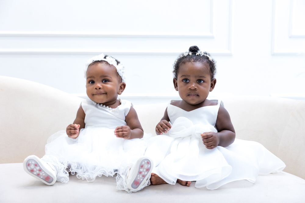 Two adorable babies wearing matching white dresses sit together on a white couch posing for a family photoshoot.