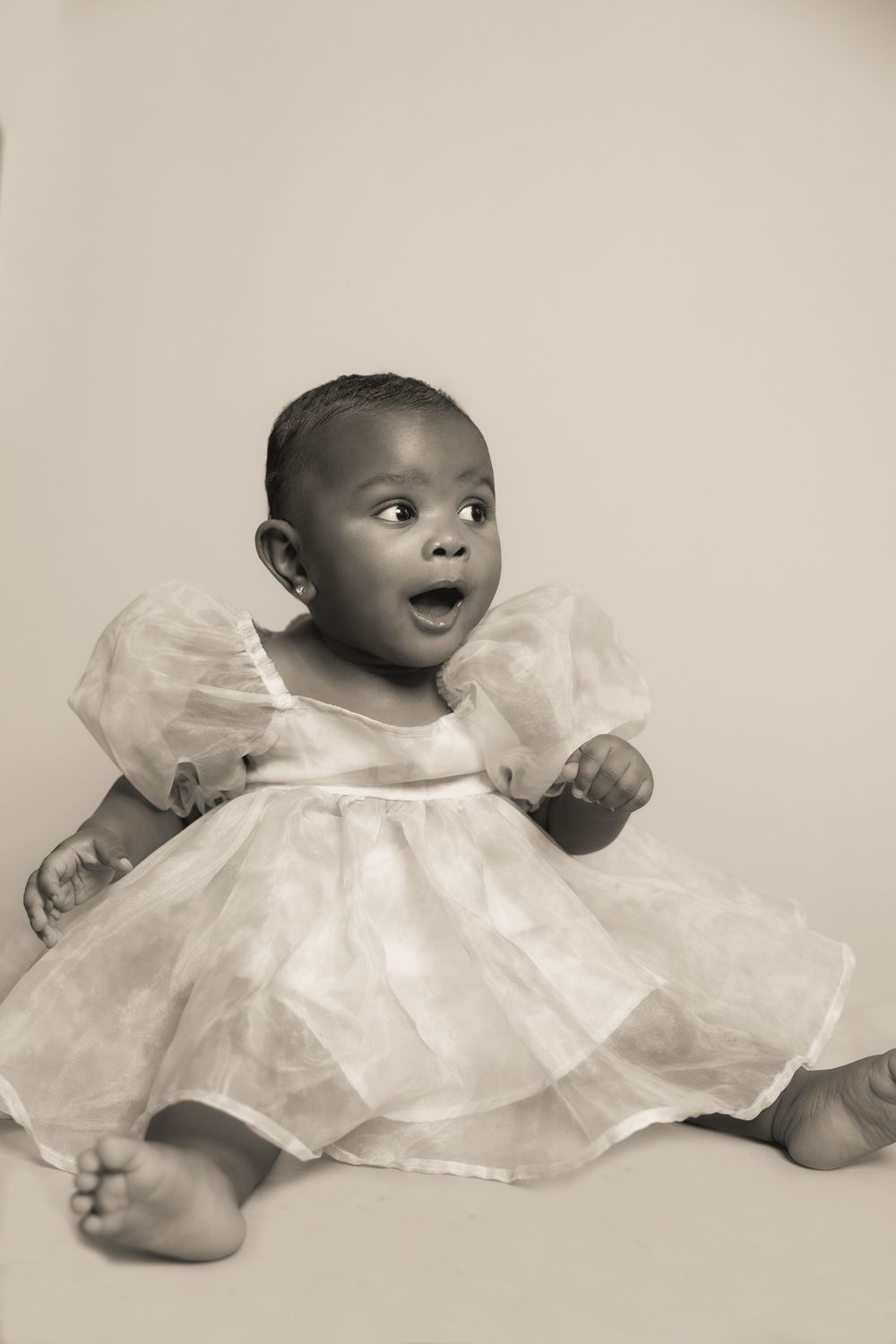 Beautiful baby in a white lacy dress sits on a light background with an expression of surprise and wonder.