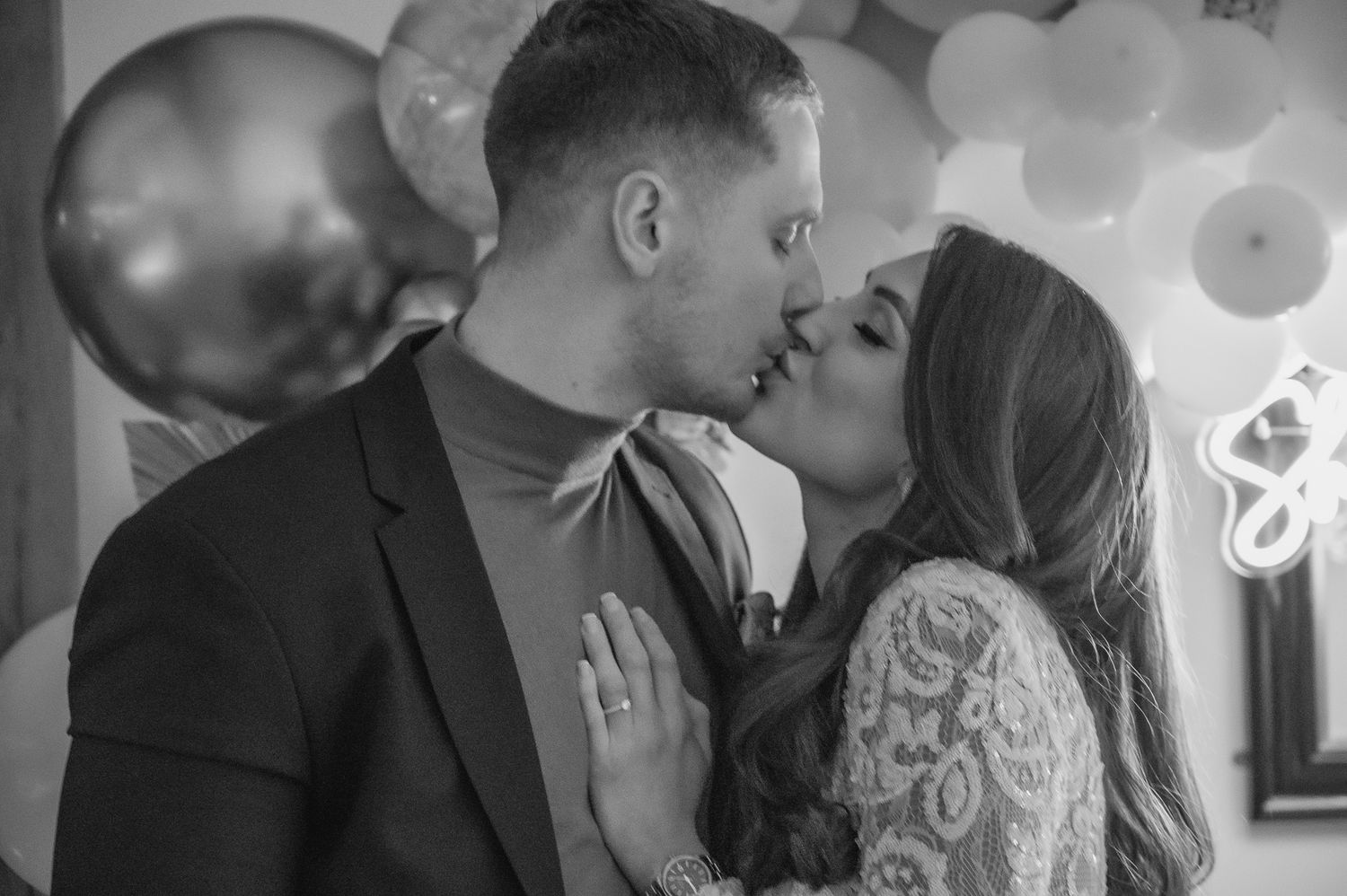 A romantic black and white portrait of a couple kissing with balloon decorations in the background.
