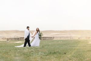 Newlyweds walk hand in hand across field at sunset with rustic benches in background.