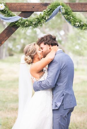 A romantic wedding kiss under a floral arch in an outdoor garden setting.