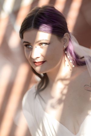 Bride in white dress with purple hair smiles in soft window lighting.