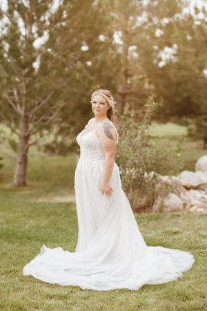 A bride in a flowing white lace gown poses gracefully on grassy grounds during an outdoor wedding photoshoot.