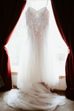 An elegant white wedding dress displayed in silhouette against window light with dramatic red curtains.