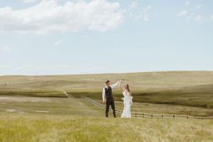 Newlyweds stand holding hands in vast prairie landscape during sunset.